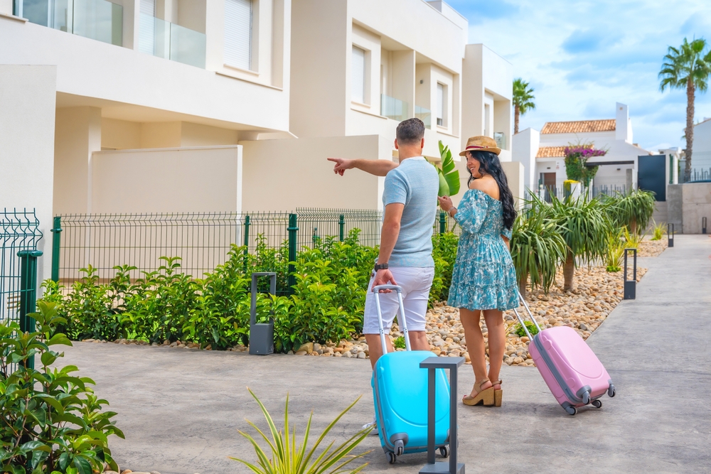 A couple with rolling suitcases arriving back at a vacation rental property, greeted by the host